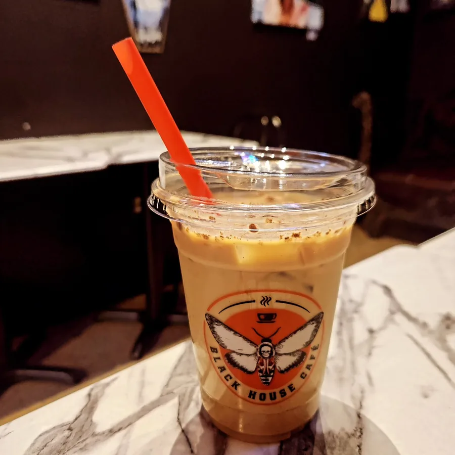 Candyman iced latte in a clear Black House Café cup with the deathshead moth logo, dusted with cinnamon and served with an orange straw on a white marble table