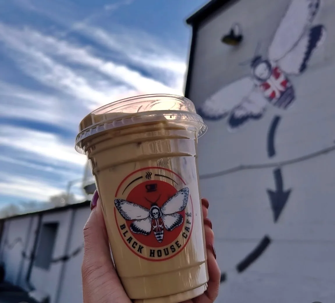 Iced latte held up in front of the death's-head moth mural on the Black House Café exterior