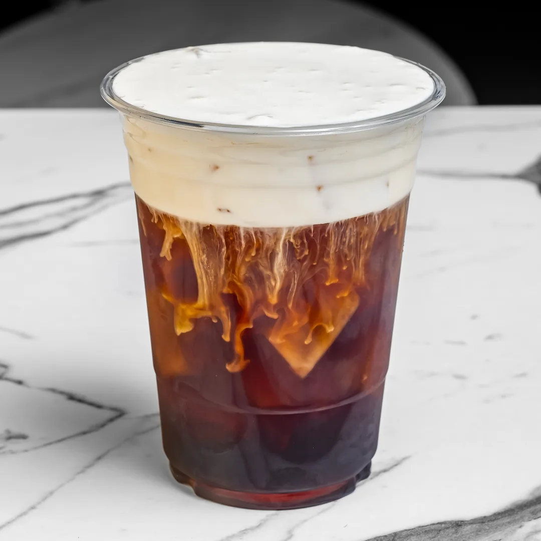 Cold brew coffee topped with cold foam in a clear plastic cup on a white marble table