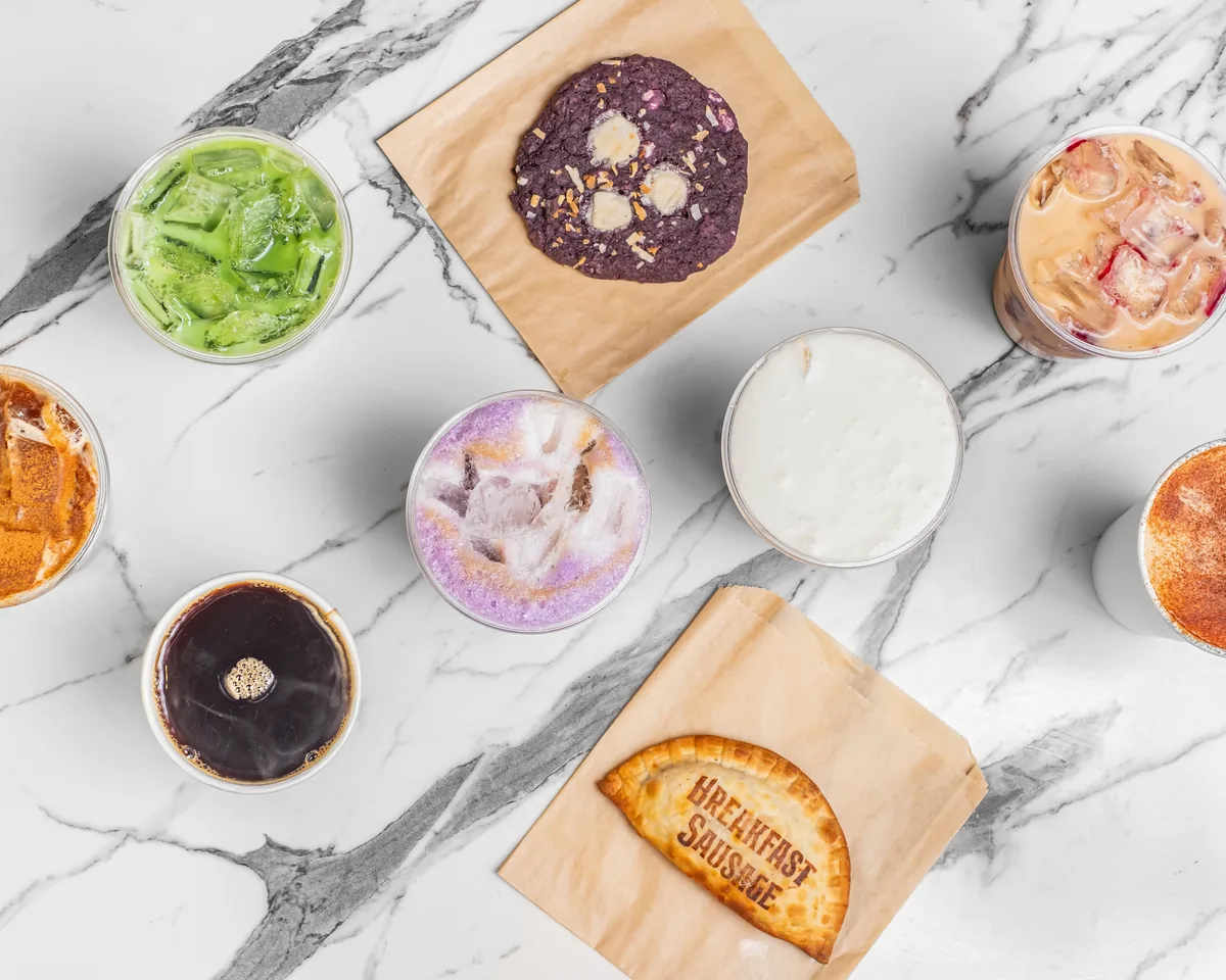 An overhead flat-lay of Black House Café iced drinks, a cookie, and a breakfast-sausage empanada arranged on a white marble table
