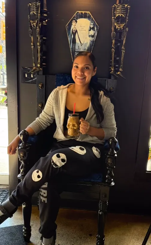 A co-owner of Black House Café seated in the café's gothic throne chair, holding a skull-shaped tumbler
